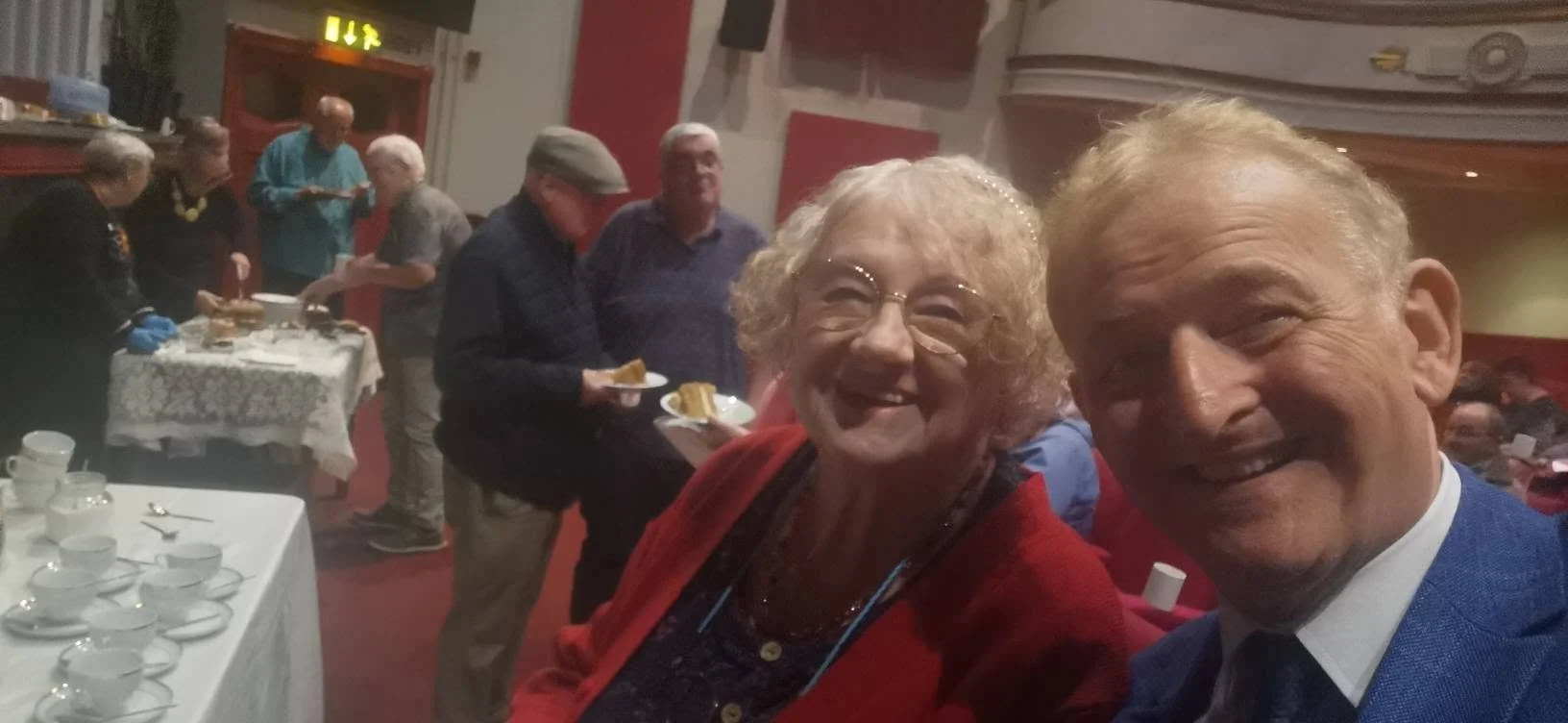 Two smiling elderly people, a woman with curly blonde hair and glasses wearing a red cardigan, and a man with blonde hair in a blue suit, taking a selfie at a social gathering or buffet event, with people serving food in the background.