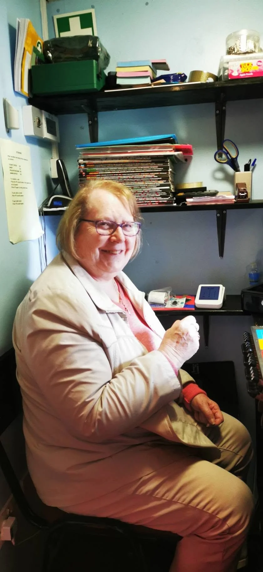 A smiling woman with glasses, wearing a tan jacket and sitting at a desk in an office with shelves filled with books, papers, and office supplies behind her.
