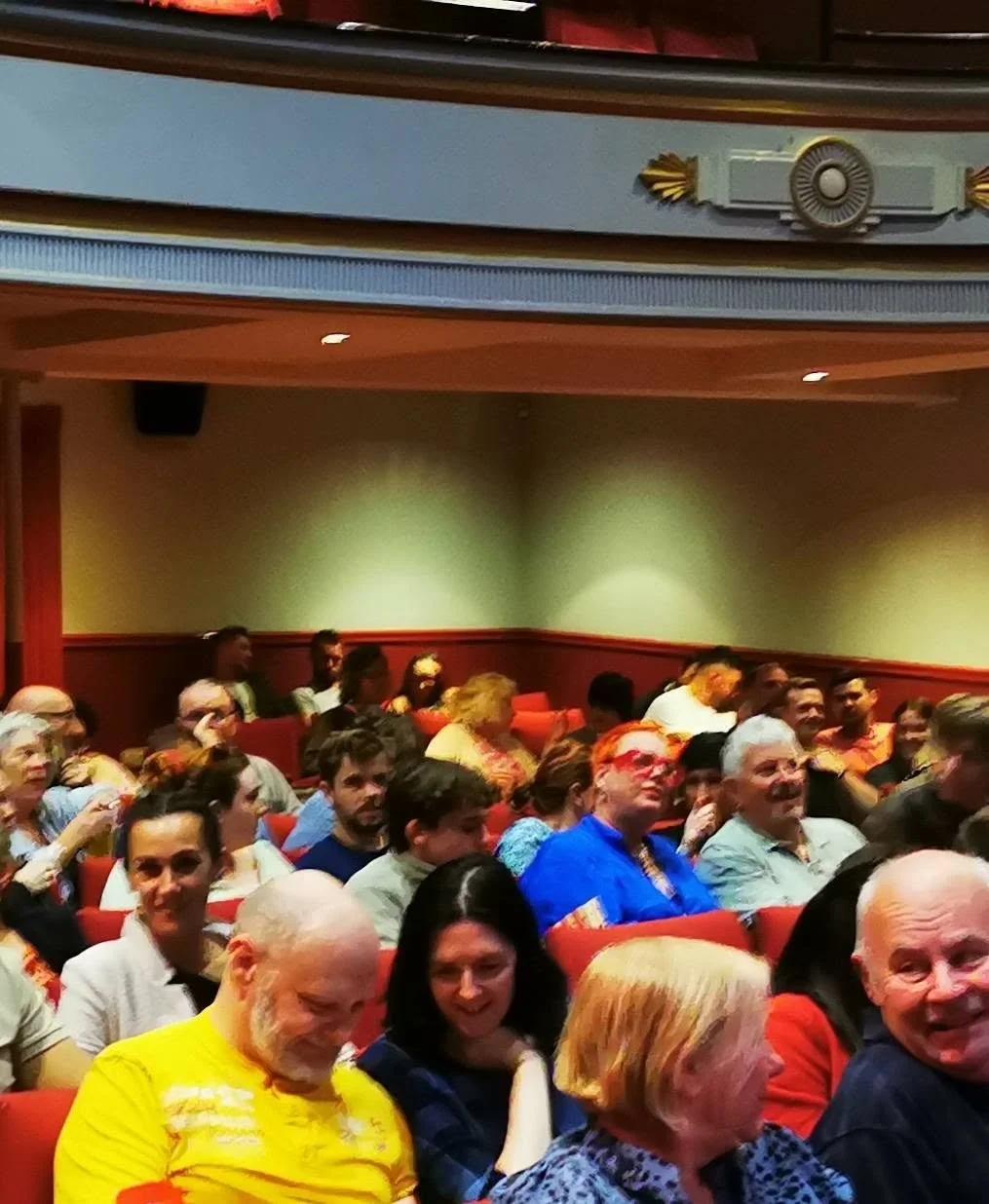 Audience seated in theater or auditorium, smiling, engaged, with diverse ages and backgrounds, red cushioned chairs, decorated walls, and stage lighting.
