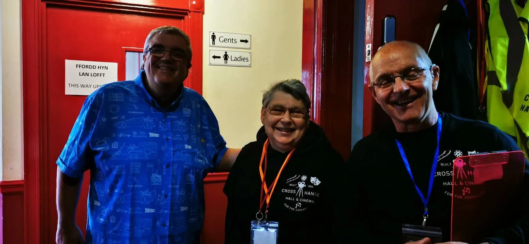 Three smiling people standing together inside a room, with signs indicating men's and women's restrooms in the background.