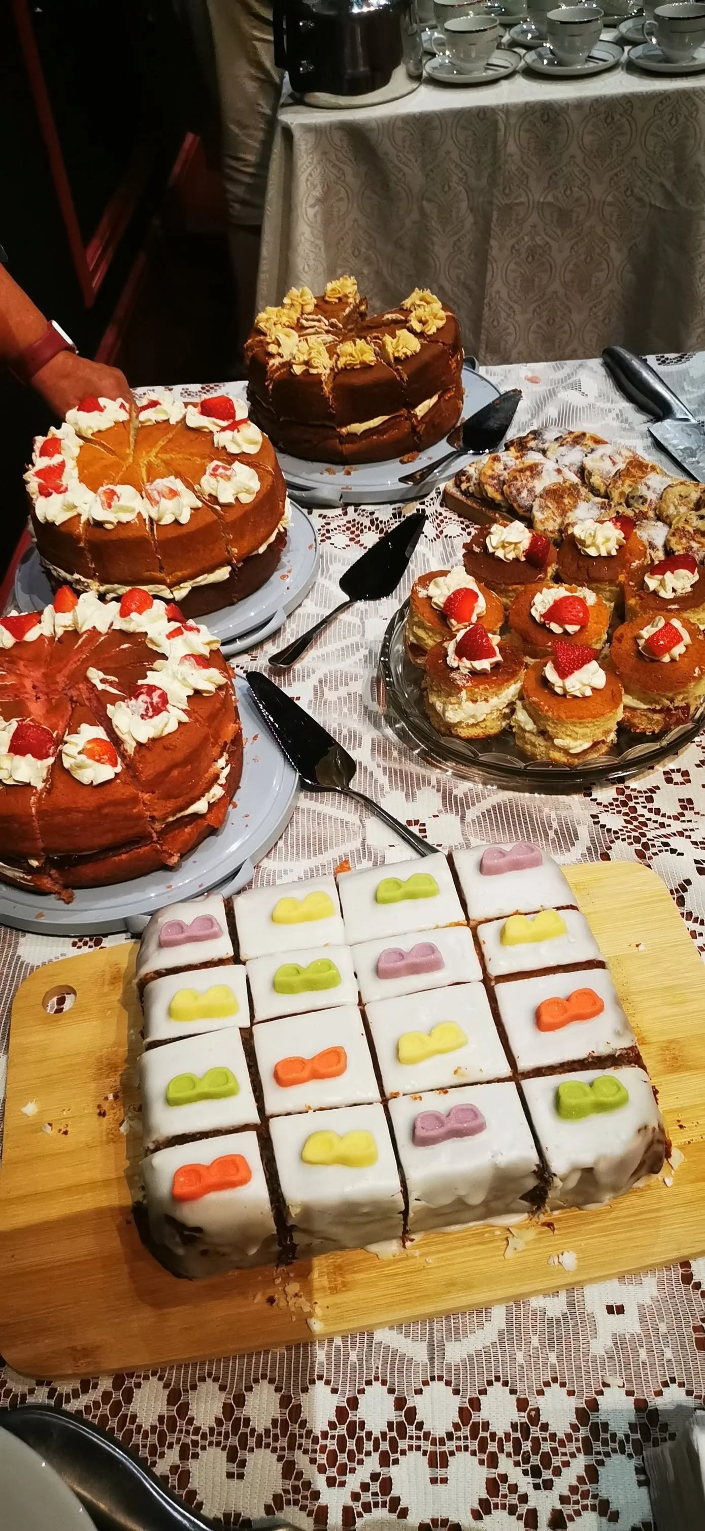 A table with an assortment of cakes and desserts, including decorated sponge cakes with whipped cream and strawberries, a chocolate cake with frosting flowers, sliced cake with powdered sugar and fruit, and a tray of small frosted square treats with 