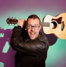 A man wearing glasses and a leather jacket smiling and holding a guitar on his shoulder against a colorful background.