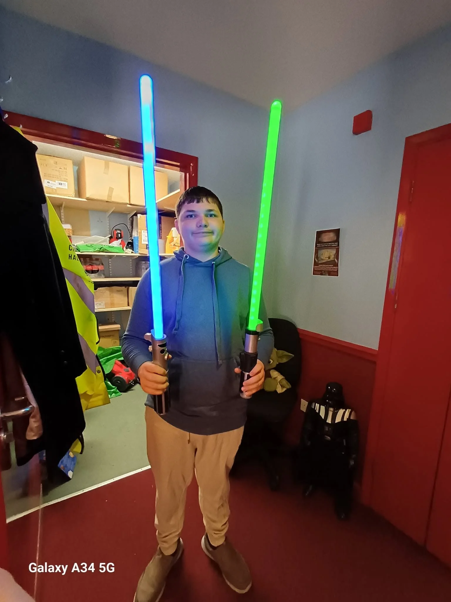 A young man holding two illuminated sticks, one blue and one green, posing inside a room with shelves and boxes in the background. The image has a Samsung Galaxy A34 5G watermark.