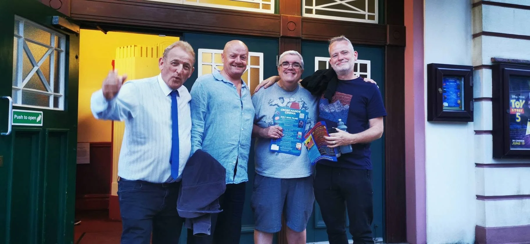 Four men smiling and standing together outside a building entrance, with posters and signage visible.