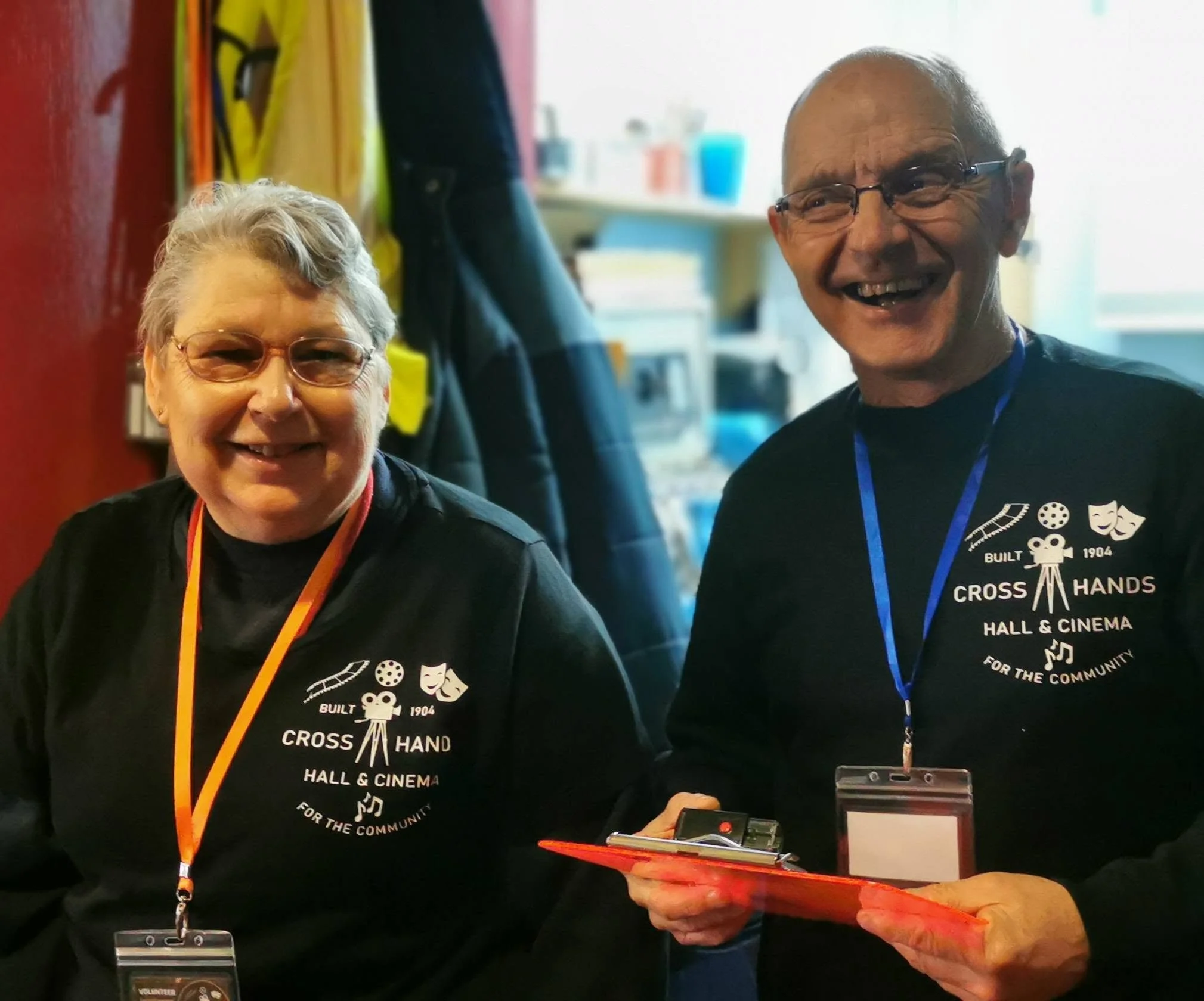 Two smiling older adults wearing black T-shirts with a logo that reads 'Built Cross Hands Hall & Cinema for the Community' at an indoor event. The woman has short gray hair and is wearing glasses, while the man has a bald head and is also wearing gla