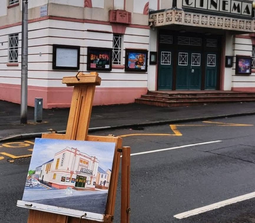 An artist's painting of a vintage theater building on an easel outside a cinema building with pink and white walls.