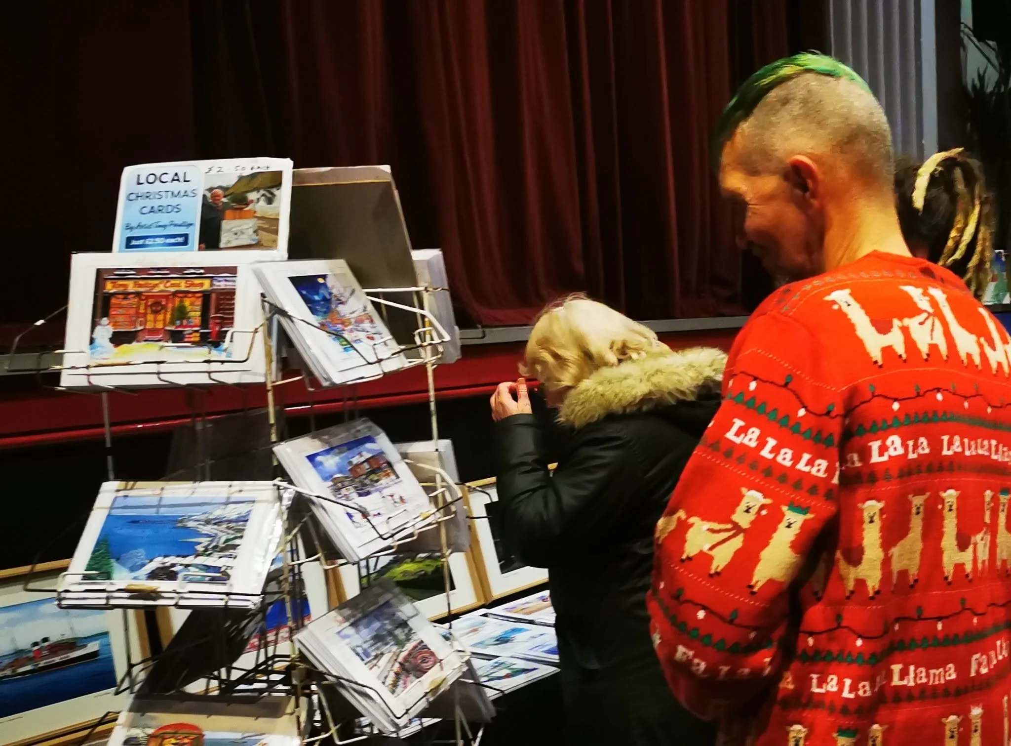 People browsing Christmas cards at a holiday market stand, with red curtains in background and colorful artwork on display.