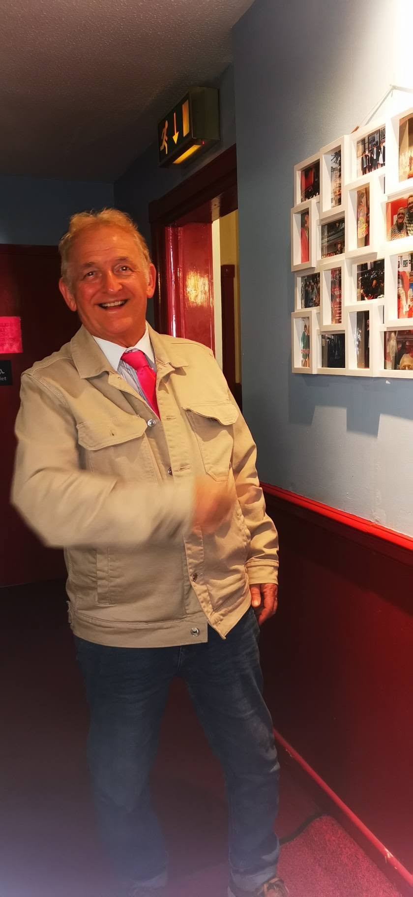 Happy man in beige jacket, white shirt, red tie, and jeans standing indoors near a wall with framed photos, smiling at the camera.