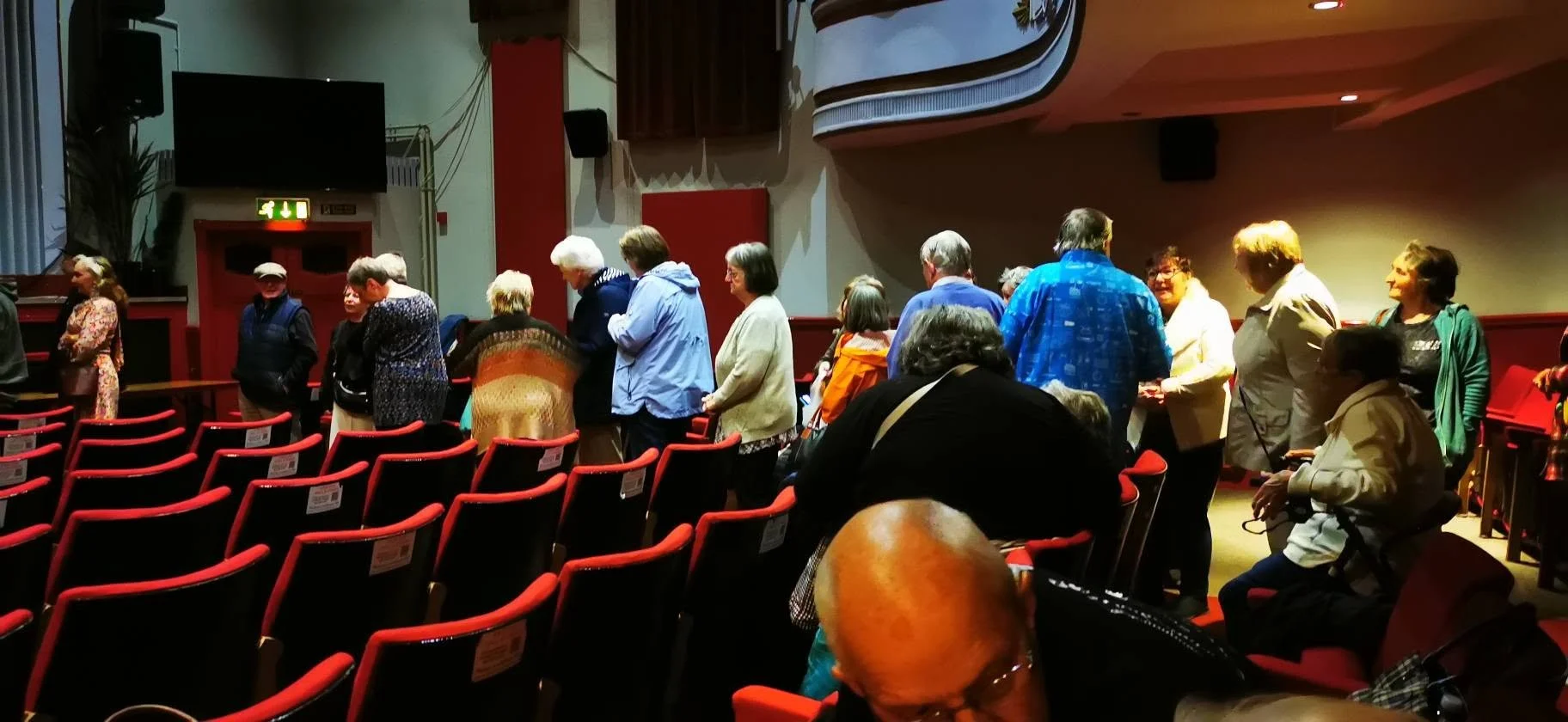 People standing in line inside an auditorium or theater with red chairs, some engaging in conversation and others waiting, under dim lighting with a screen and vent on the wall.