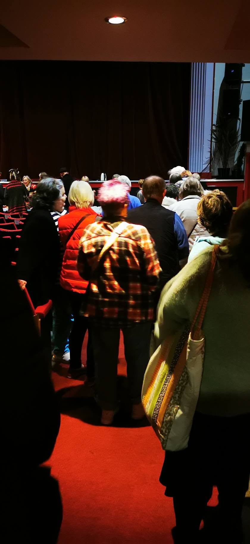 Audience members waiting inside a theater or auditorium, seated and standing near a stage with a closed curtain.