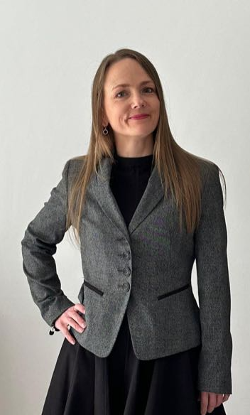A woman with long brown hair, wearing a gray blazer over a black top, standing against a plain white background.