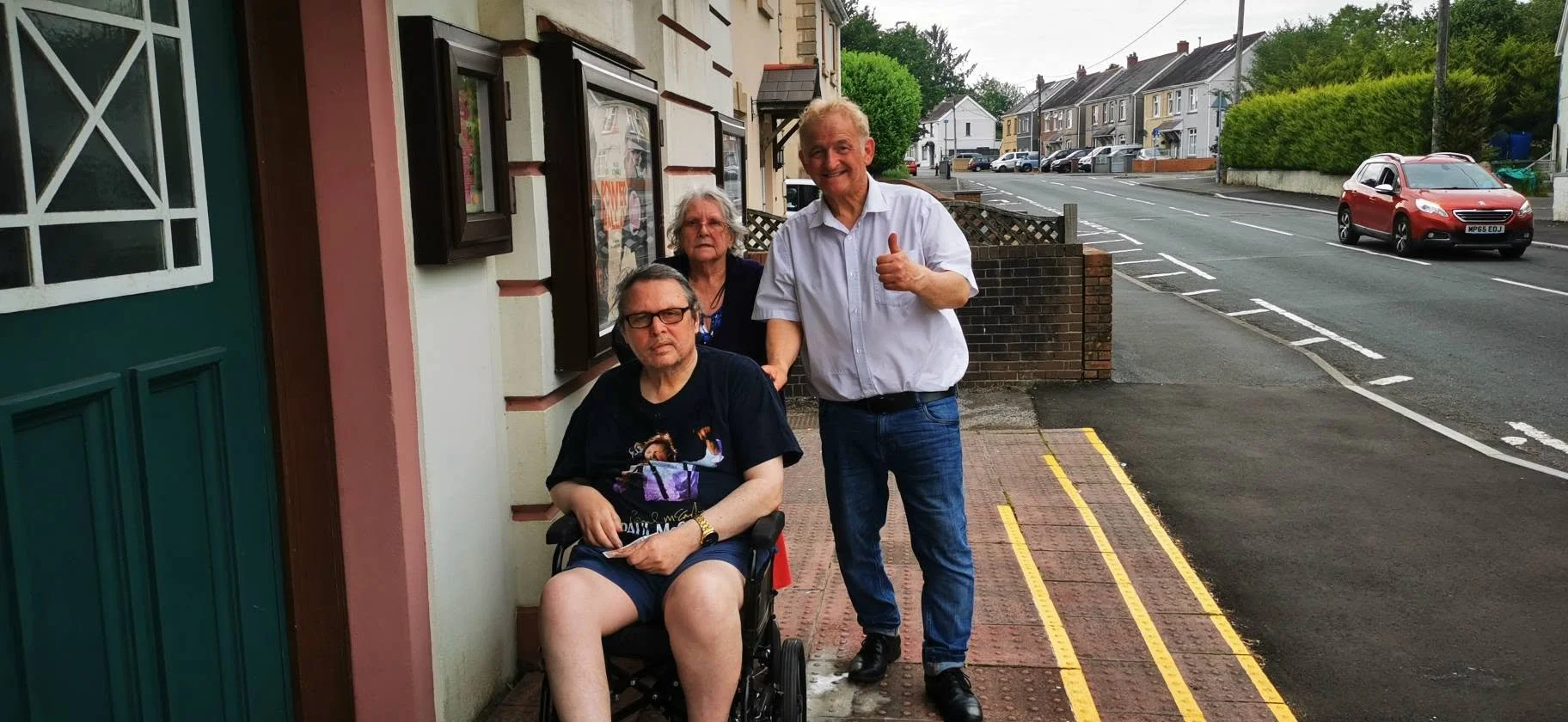 Three people on a sidewalk in a suburban neighborhood. One person is seated in a wheelchair, wearing glasses and a black T-shirt, holding a phone. A man in a white shirt and jeans stands next to them, giving a thumbs up. Another woman with gray hair 