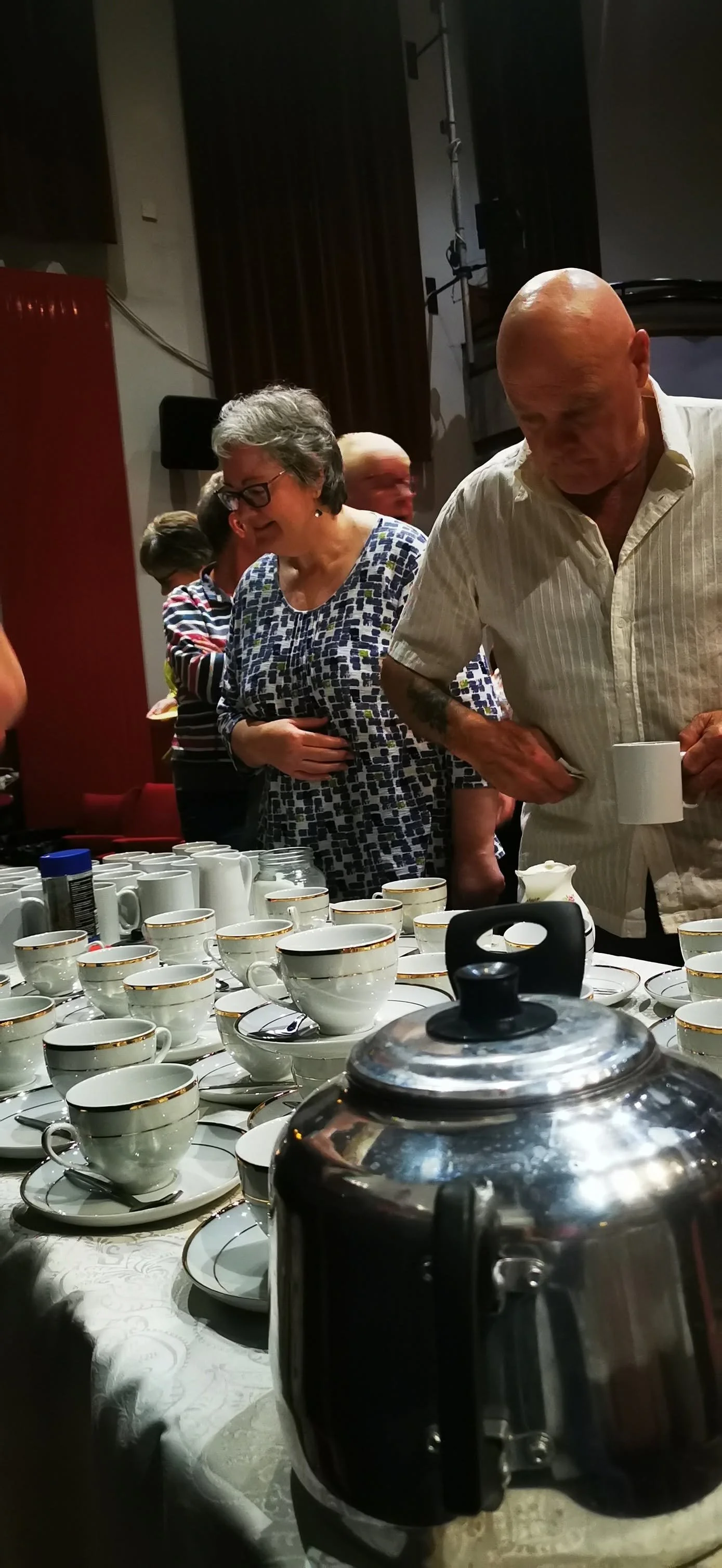 People preparing and serving tea at a gathering with multiple cups, a coffee pot, and cups on a table.