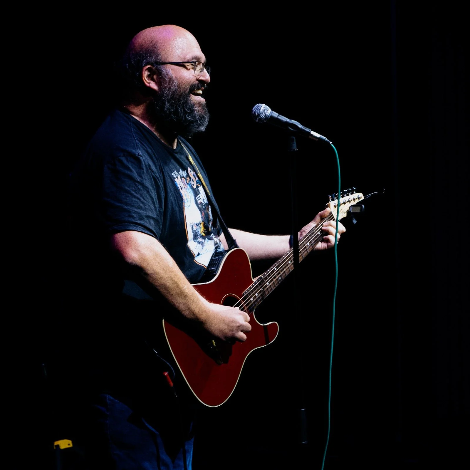 Man with a beard and glasses singing and playing an acoustic guitar on stage with a microphone.