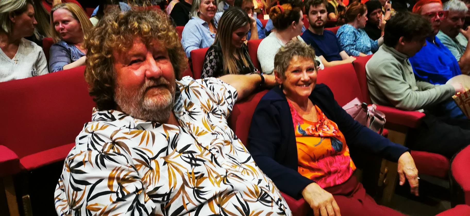A group of diverse people sitting in a theater, with two smiling women and a bearded man in the foreground.