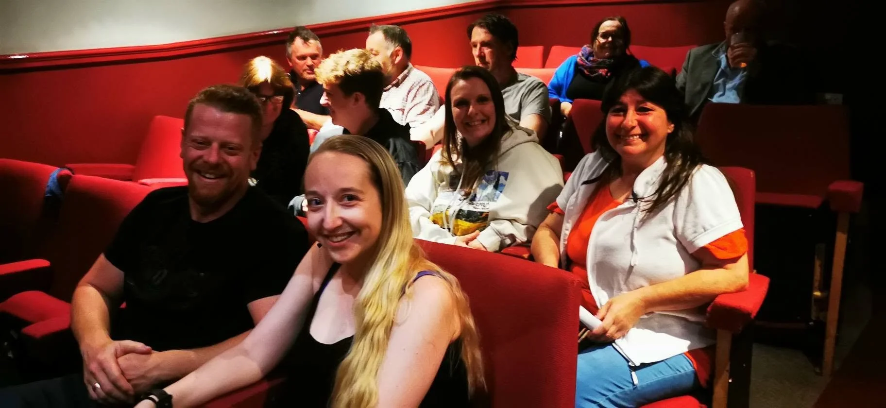 Group of people sitting in red theater seats, smiling, in a dimly lit venue.