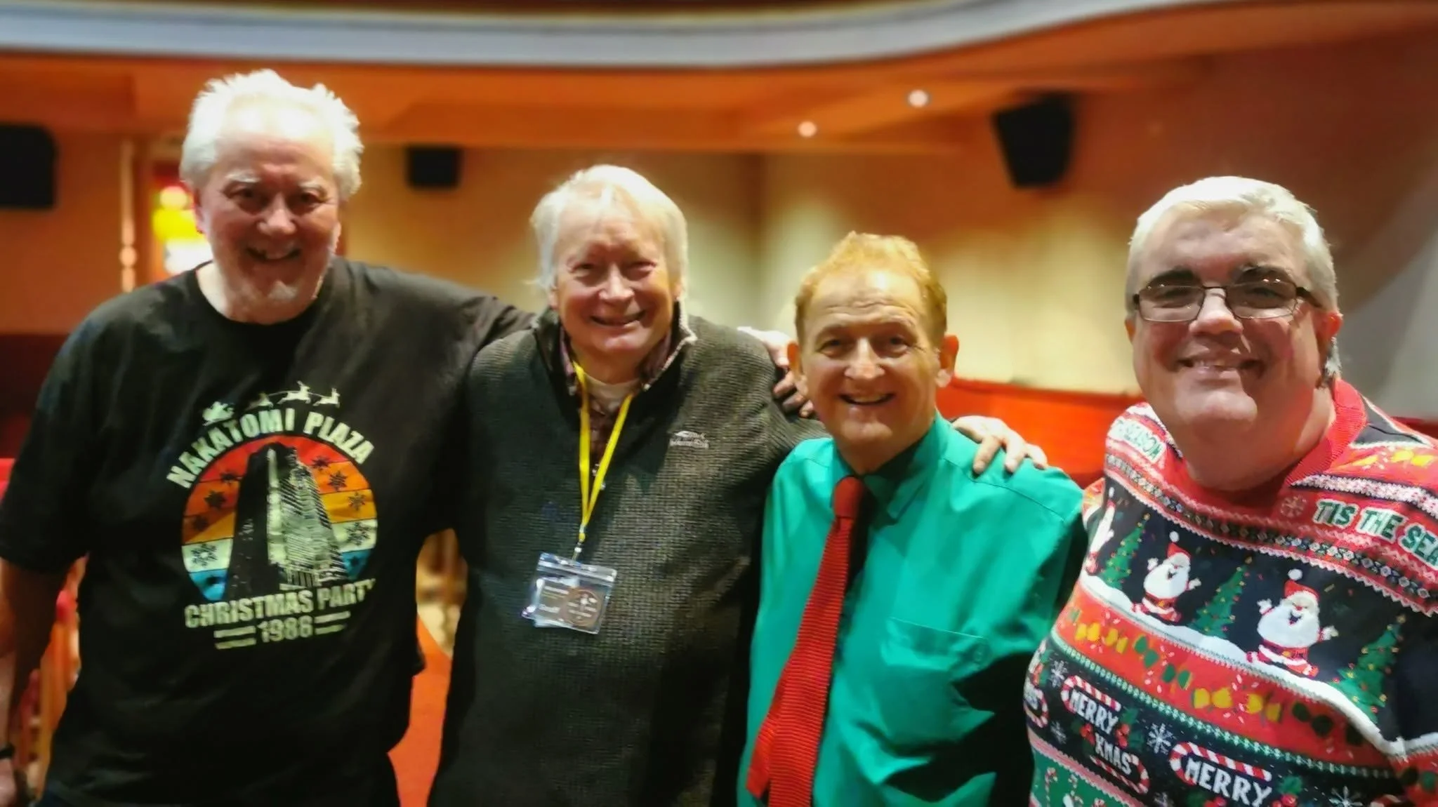 Four men smiling and posing together indoors, three of them wearing colorful shirts and ties, one wearing a festive Christmas sweater, celebrating at a holiday gathering.