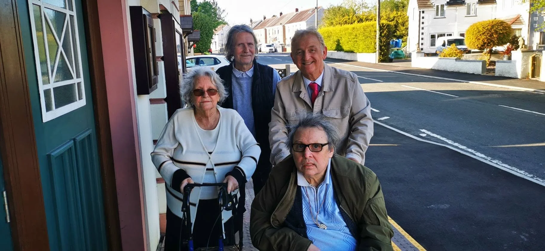 Five senior individuals, three men and two women, standing and sitting outside on a sidewalk in a suburban neighborhood. The elderly woman is using a walker, and they are all smiling or looking at the camera. The background includes houses, trees, an