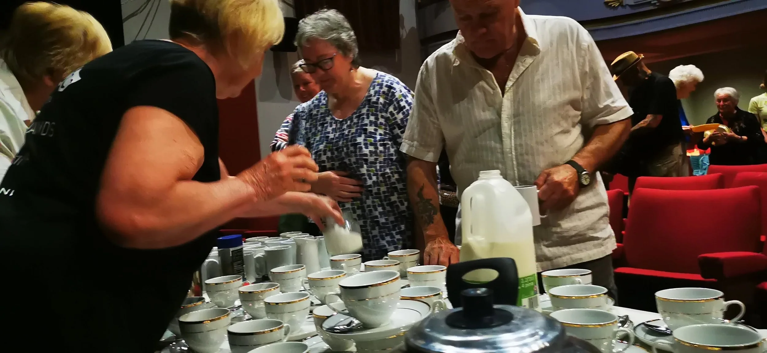 People gathered around a table with cups, preparing or serving beverages, in a room with red chairs and a group of individuals in the background.
