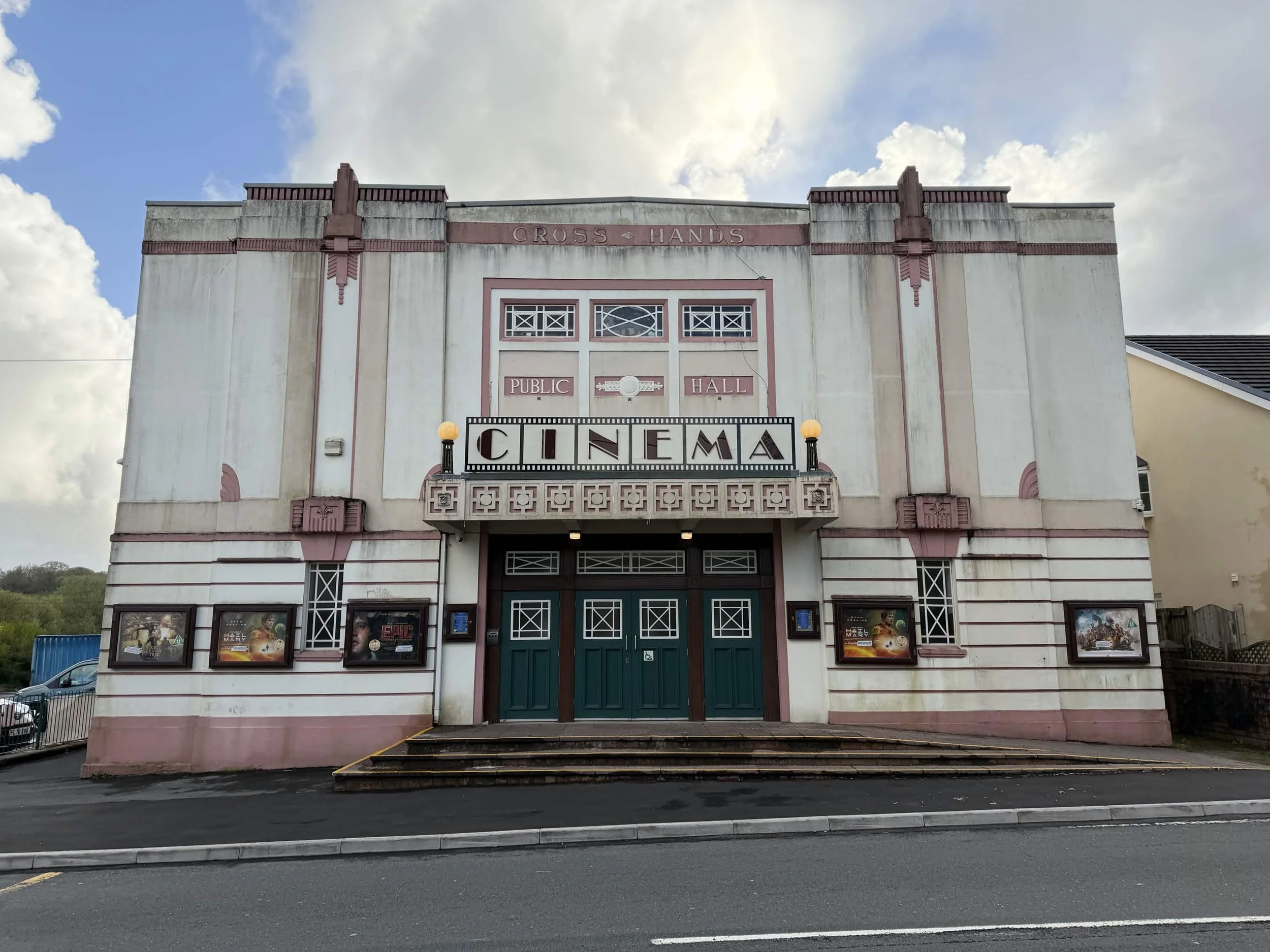 Front view of a vintage-style community theater building with a marquee that says 'CINEMA,' and above it, a sign that reads 'CROSS HANDS PUBLIC HALL,' with posters displayed on either side of the entrance and steps leading up to the doors.