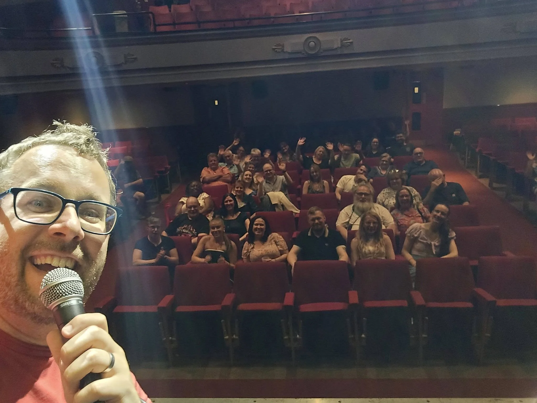 A man with glasses and a beard holding a microphone, taking a selfie in front of an audience seated in a theater with red seats, some of whom are waving at the camera.