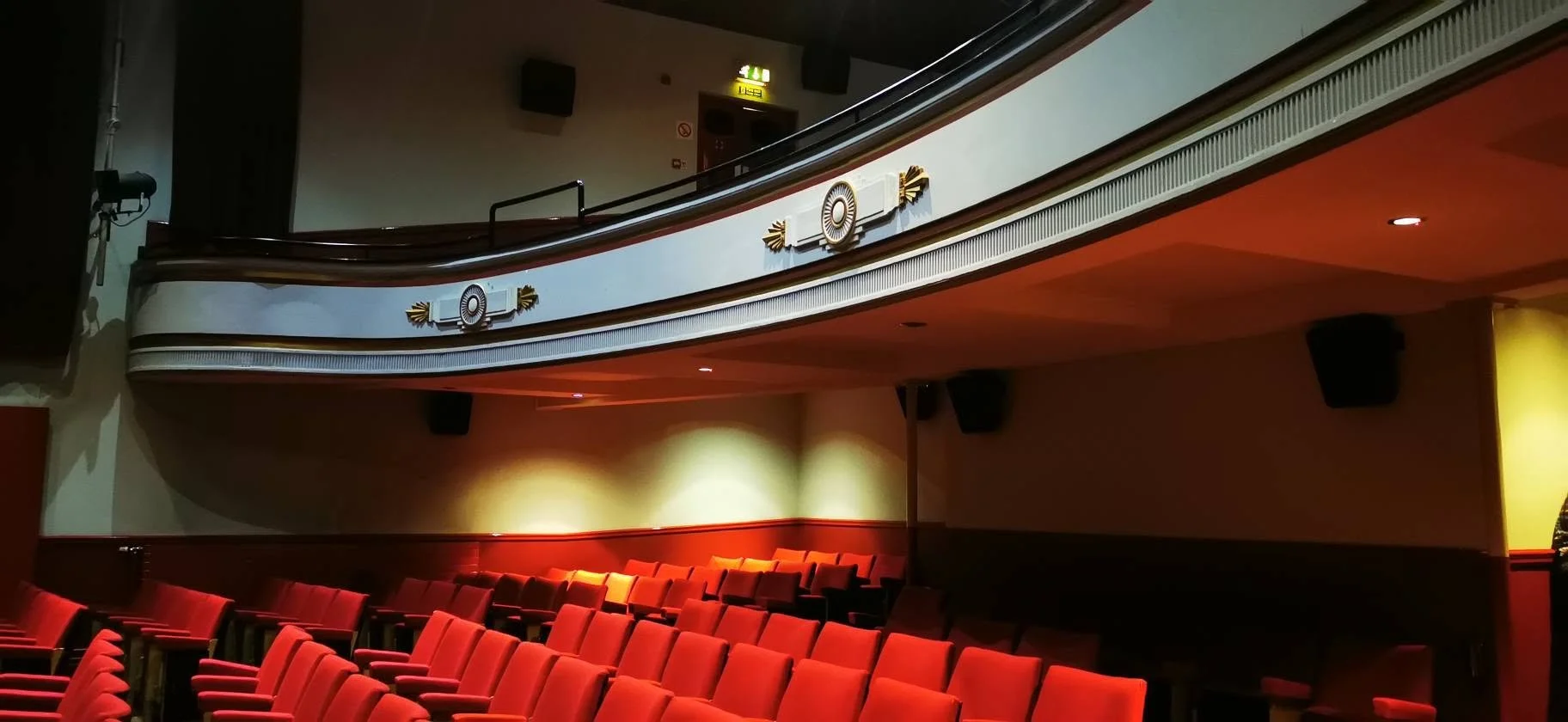 Interior of a theater with red chairs on the ground floor and a balcony above, decorated with ornate gold and white accents, and large black speakers mounted on the walls.