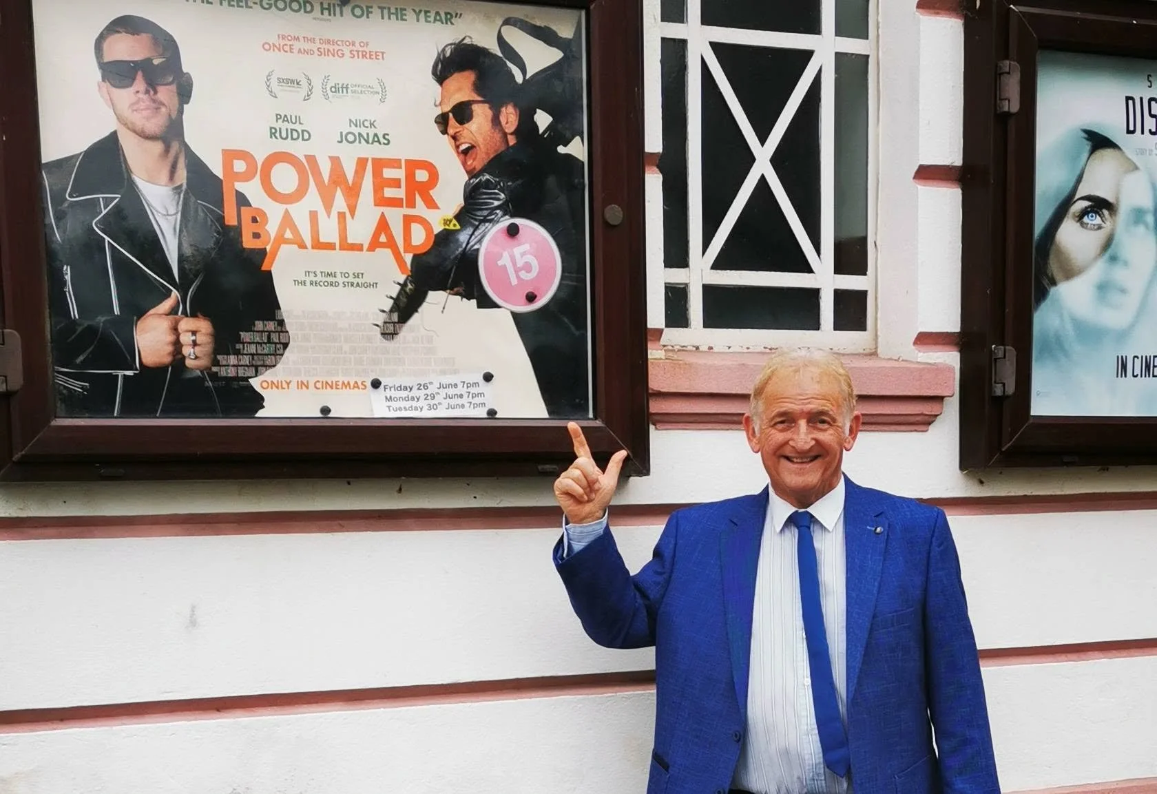 An older man in a blue suit smiling and pointing at a movie poster for 'Power Ballad' on a wall.