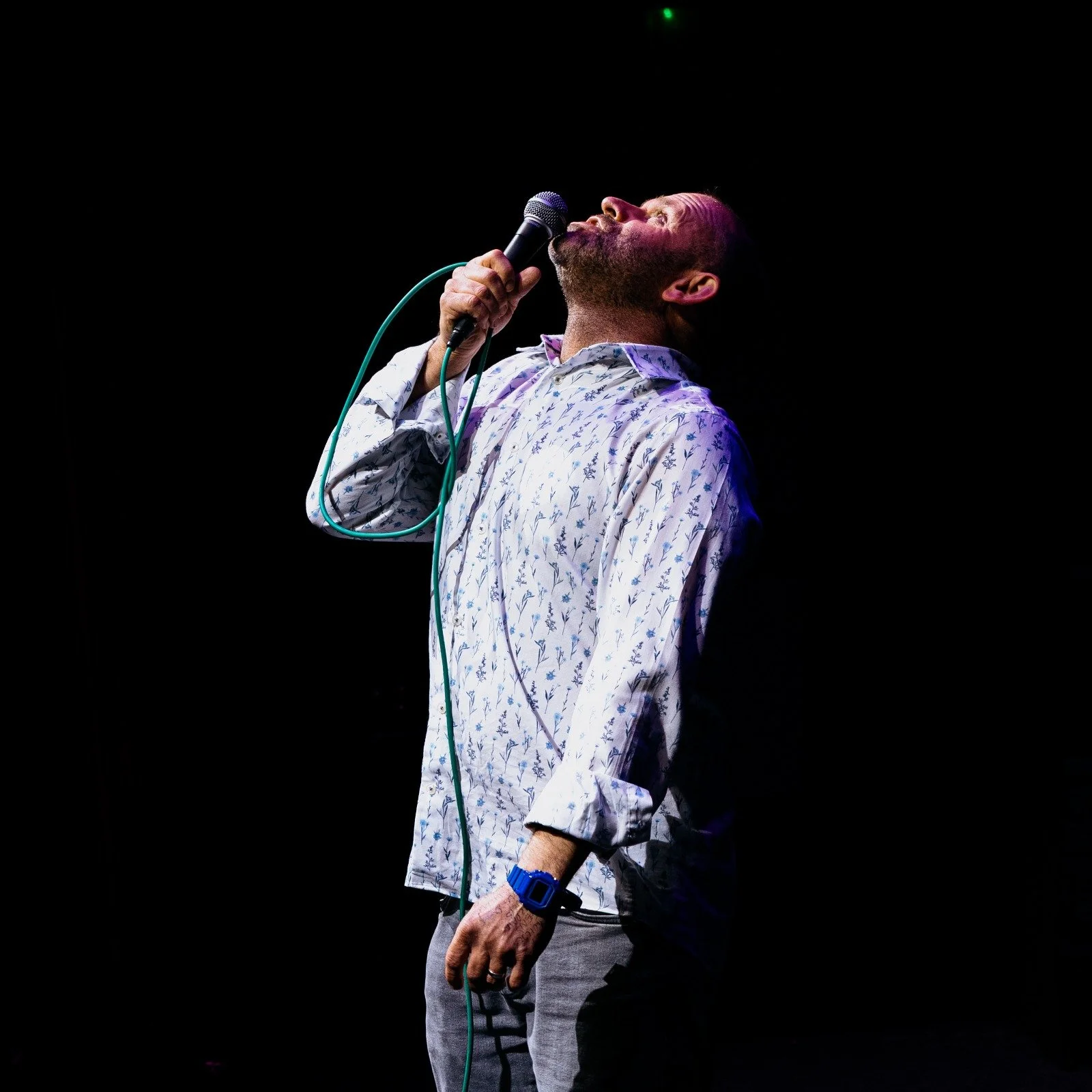 Man singing into a microphone on stage, wearing a patterned button-up shirt and gray pants, with a black background.