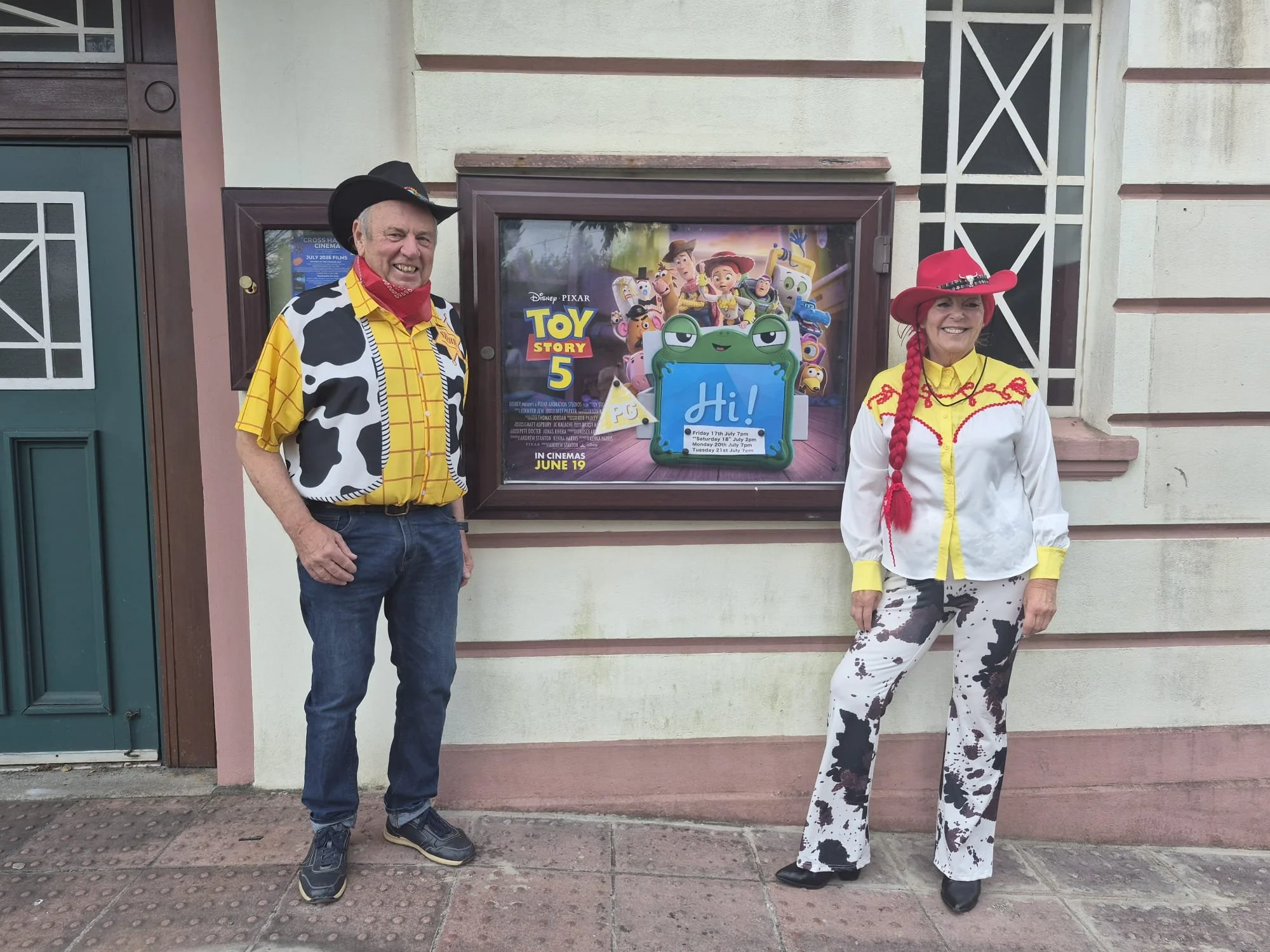 Two people dressed as cowboys standing outside in front of a movie poster for Toy Story 5.