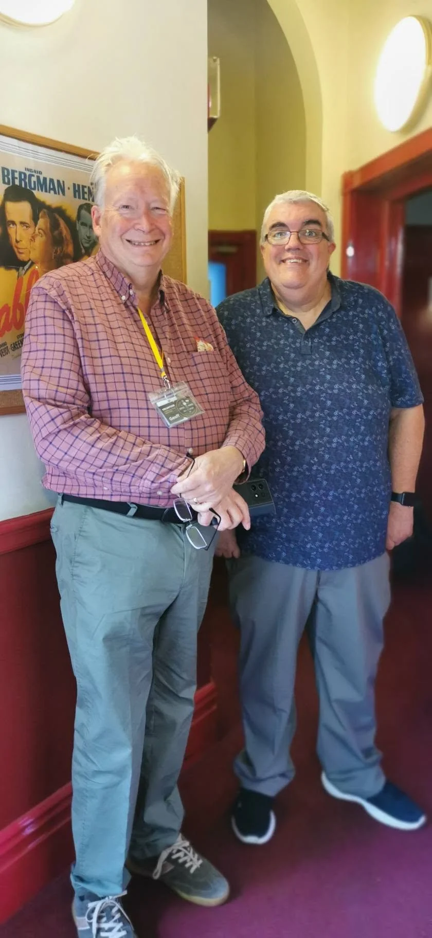 Two smiling men standing indoors, one with glasses and a badge, the other wearing a patterned shirt, with a poster on the wall behind them.