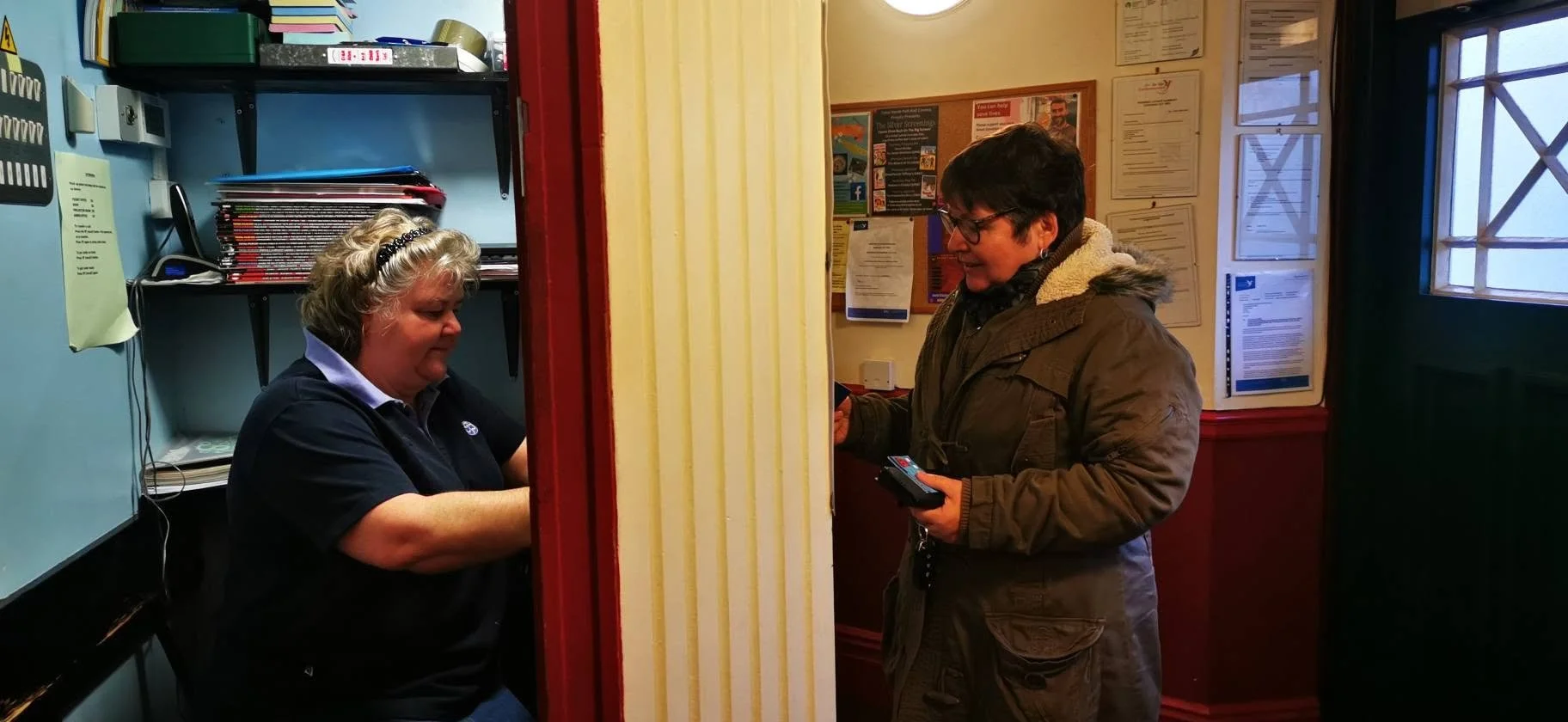 A woman at a counter receiving service from another woman in a small room. The woman at the counter is holding a phone and smiling, while the woman behind the counter is sitting and looking down at something.