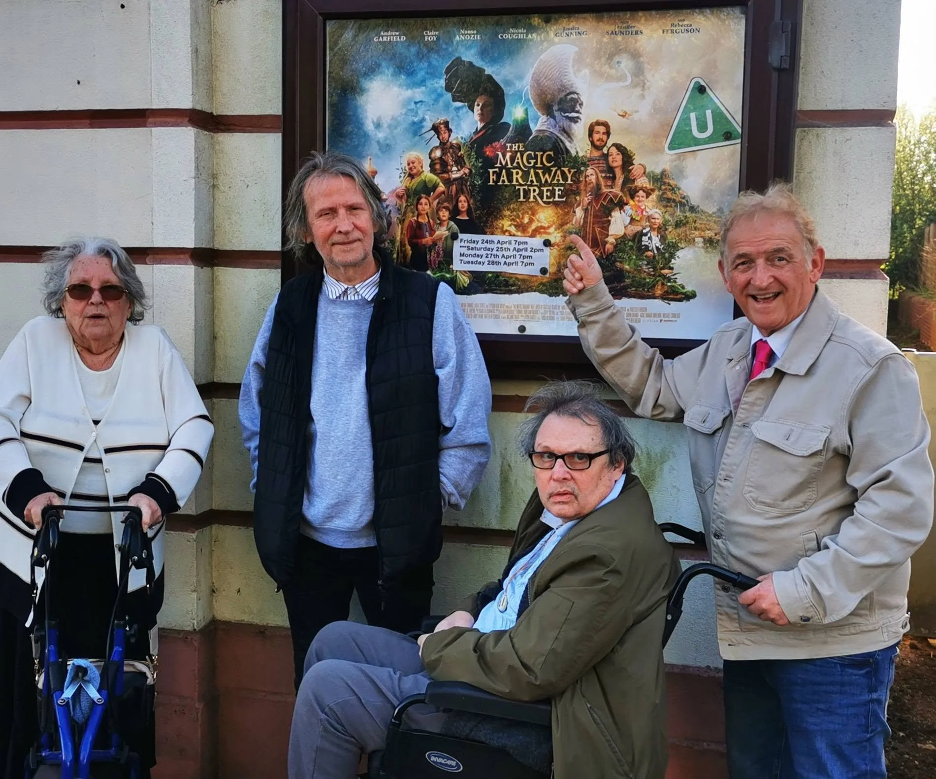 Four elderly individuals, three men and one woman, pose in front of a movie poster for "The Magic Faraway Tree." The woman is using a walker, and one man is sitting in a wheelchair. The other two men are standing, with one pointing at the poster. All