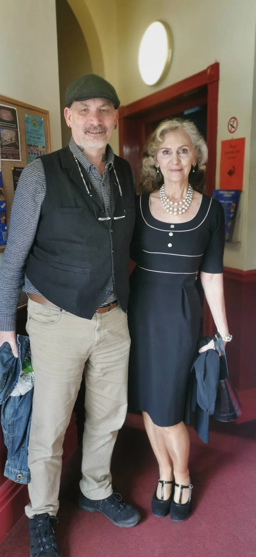 A man with gray hair and beard wearing a black cap, black vest, and beige pants standing next to a woman with curly gray hair in a black vintage-style dress with pearl necklace, standing inside a building near a doorway.