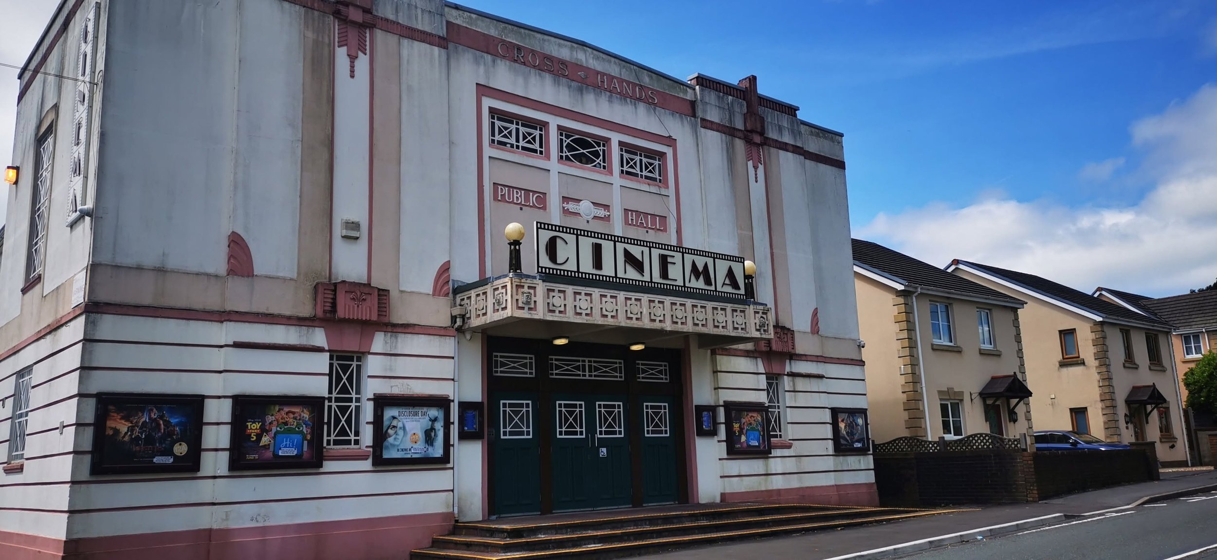 The exterior of an old-fashioned cinema building with a large sign reading 'CINEMA' and decorative architectural elements, situated in a suburban area with houses nearby.