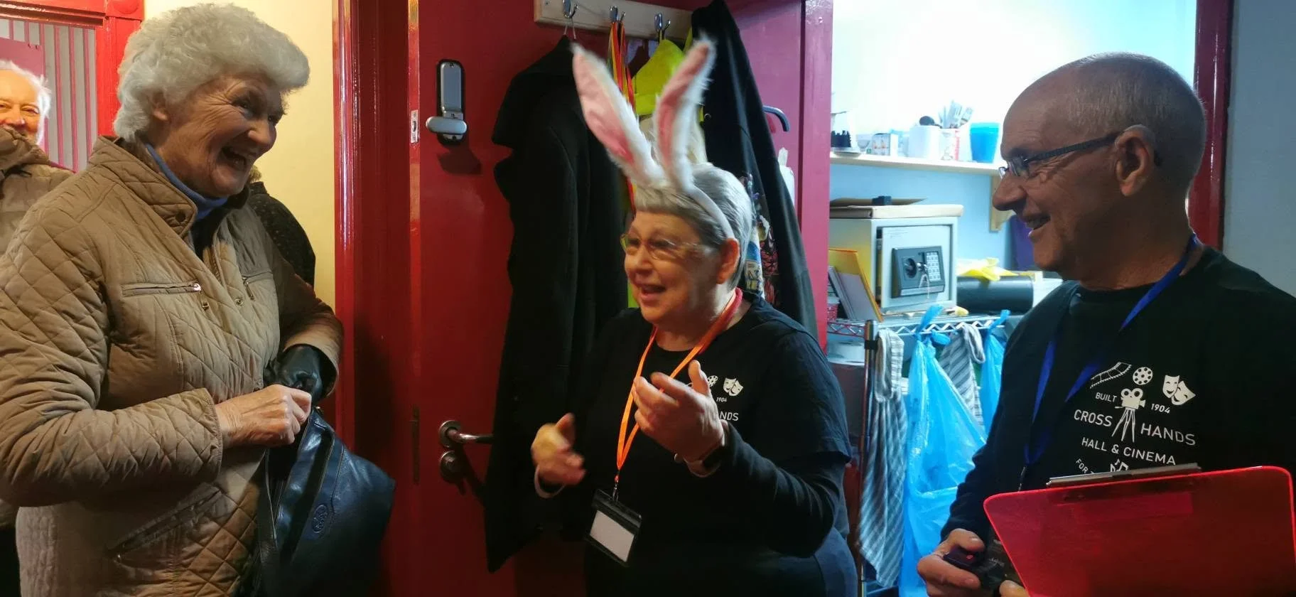 A group of three people inside a room, engaging in conversation. One woman is wearing novelty bunny ears and glasses, smiling and gesturing with her hand. The other woman, with gray hair, clad in a quilted jacket, is smiling back. A man, with glasses