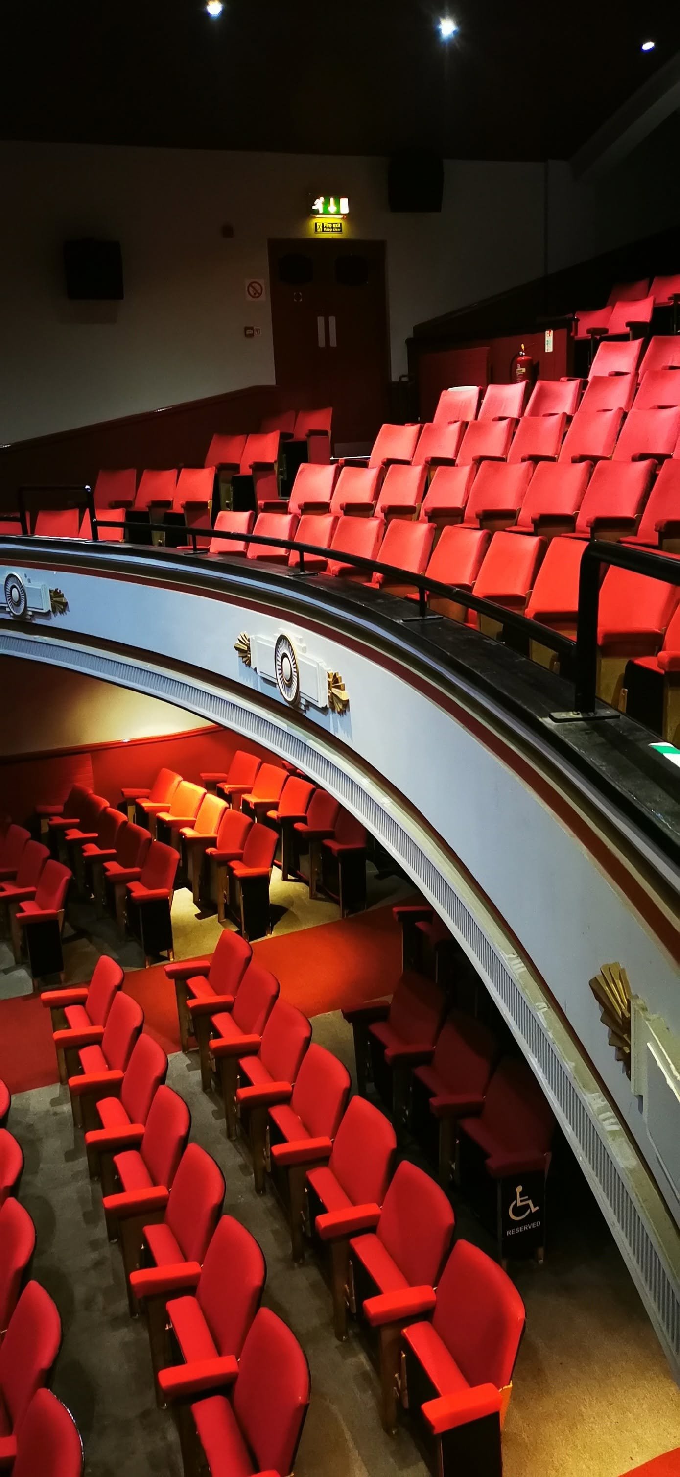 Empty theater with red seats arranged in multiple tiers and a decorative balcony railing.