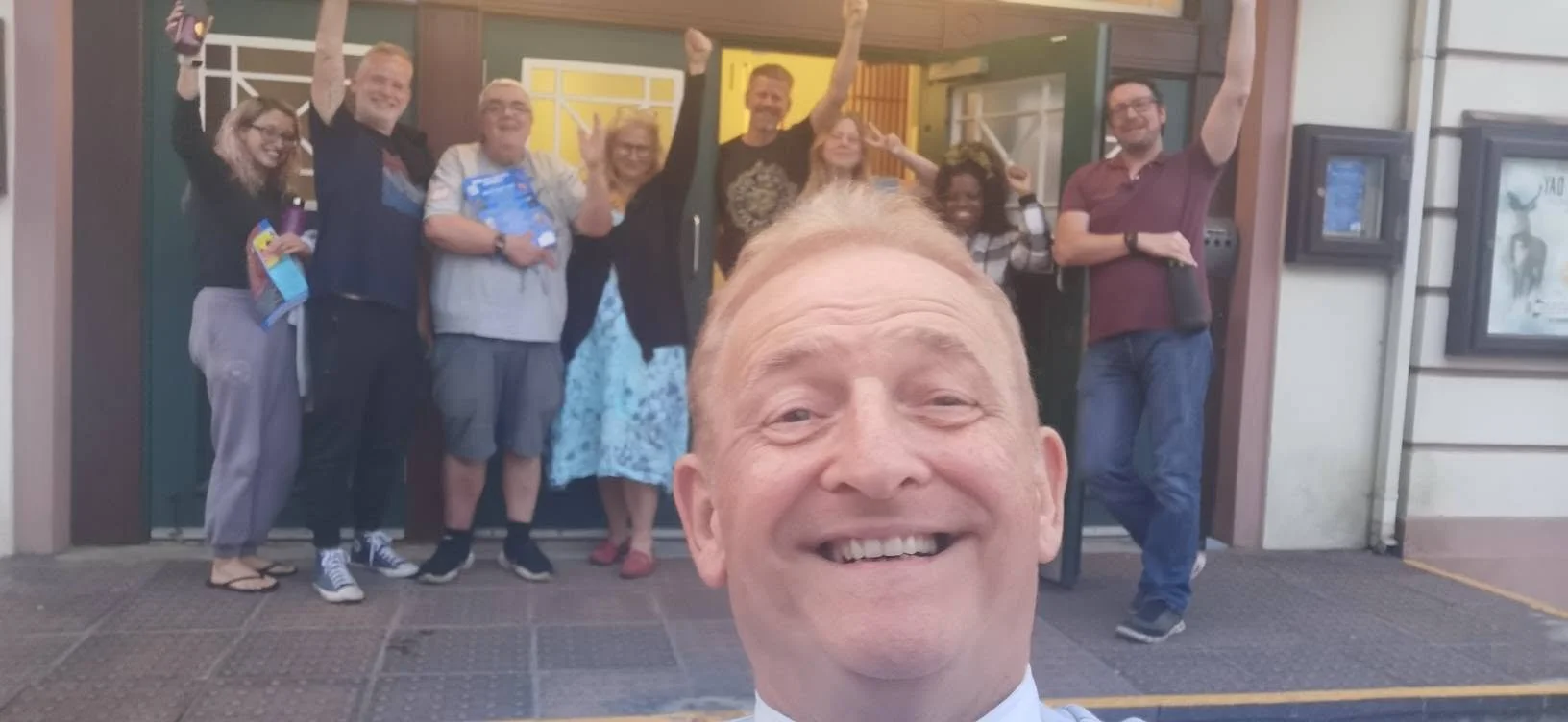 A group of nine people standing in front of a colorful building, shaking hands, smiling, and making peace signs. A man in the foreground taking a selfie.