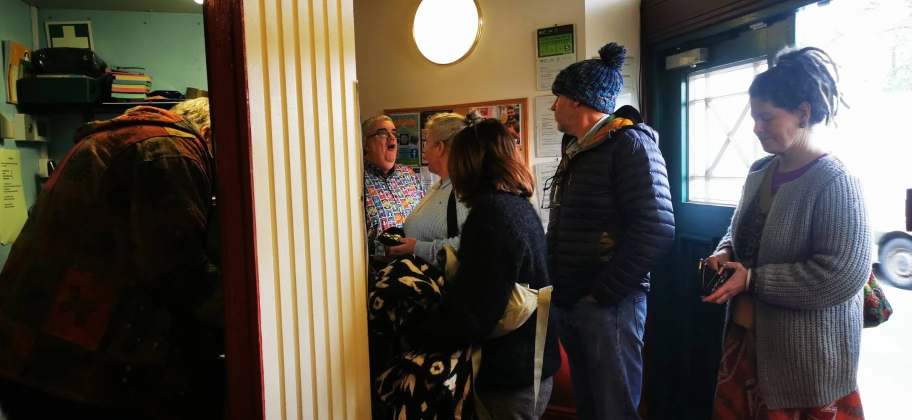 People standing in line at a small check-in counter of a cafe or restaurant, with a woman inside the counter talking to a woman and others waiting in line.