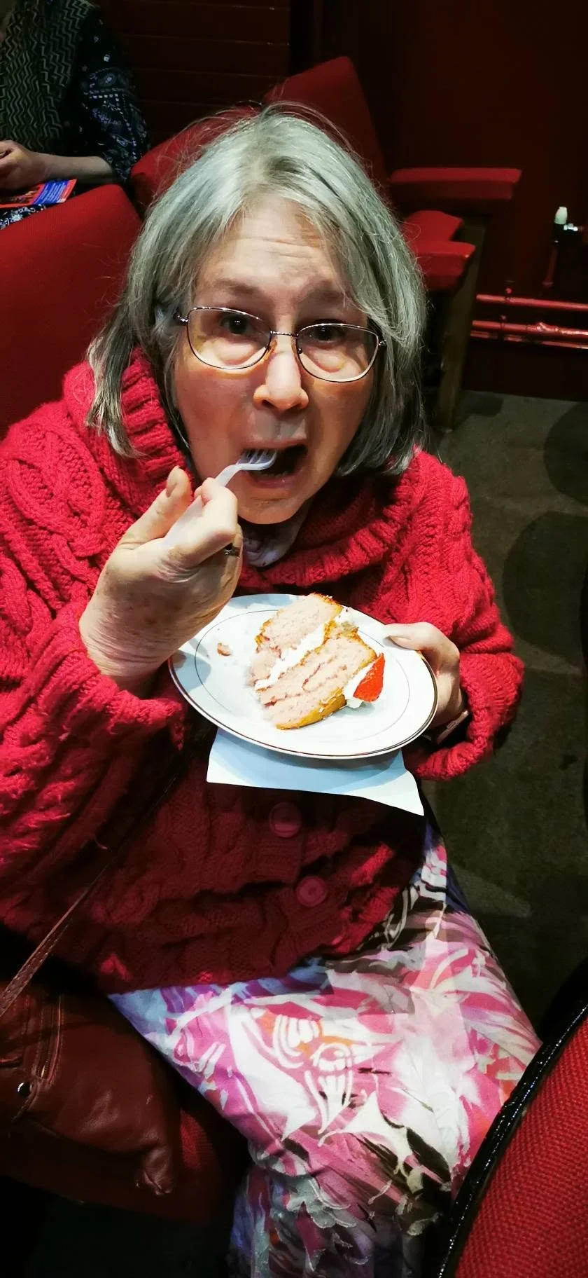An older woman with glasses wearing a red knitted sweater sitting in a restaurant, holding a plate with a slice of cake, using a fork to eat.