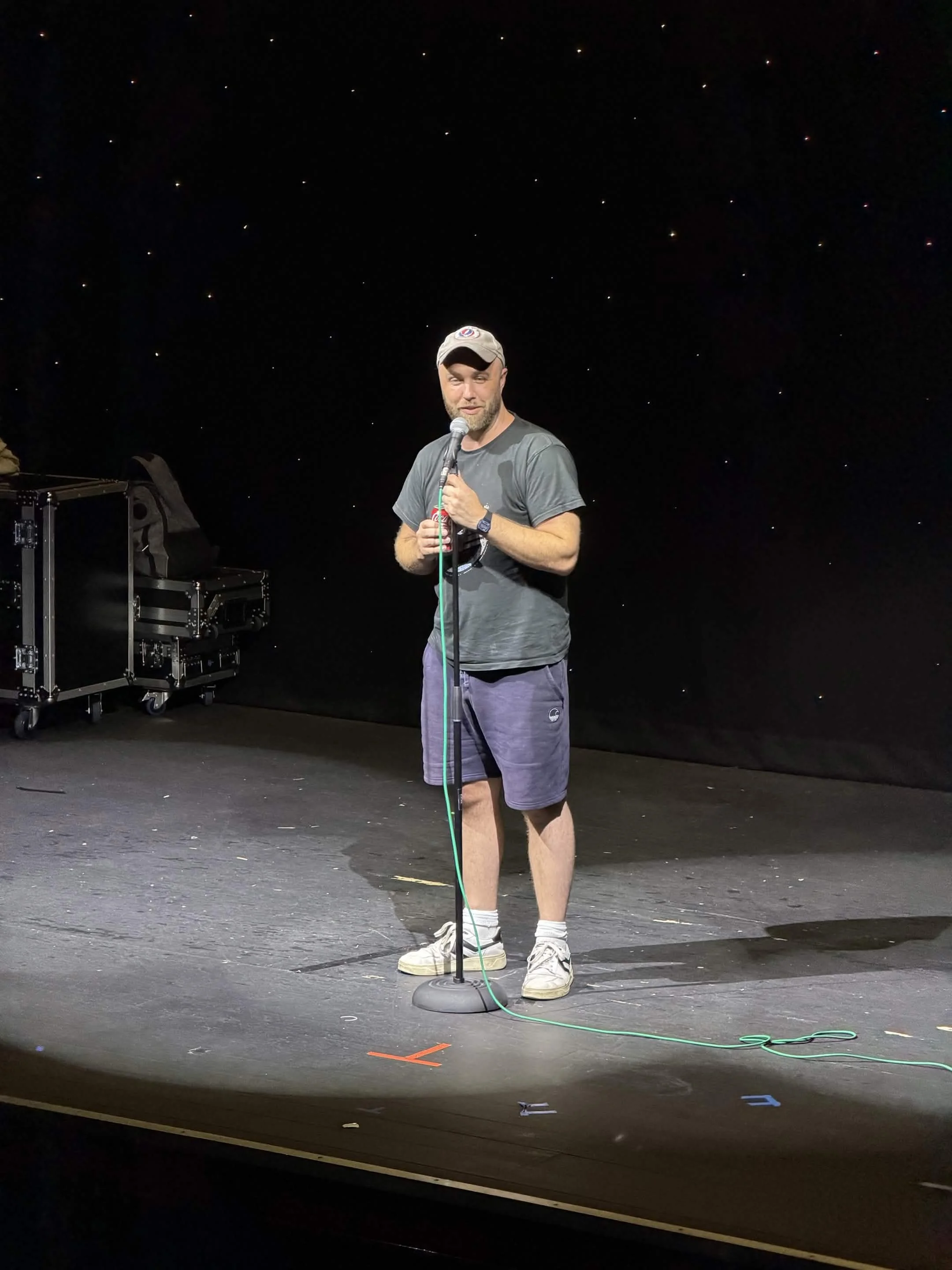 A man standing on a stage with a black starry background, holding a microphone, wearing a gray t-shirt, purple shorts, and a beige cap, with equipment and a green cable on the stage.