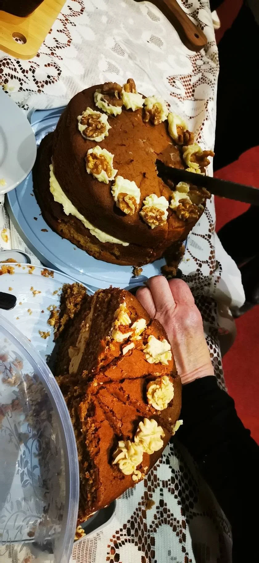 A person holding a slice of pumpkin cake with cream and walnuts on top, next to a larger pumpkin-shaped cake decorated similarly, both on a table with a lace tablecloth.