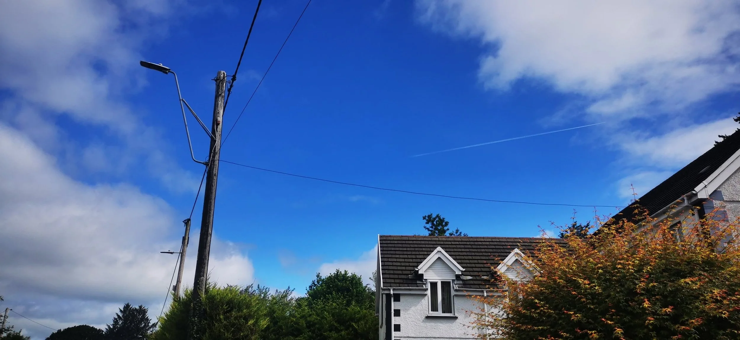 A wooden utility pole with a streetlight, power lines, a house with a gray roof, white walls, and a small attic window, trees, and a blue sky with white clouds.
