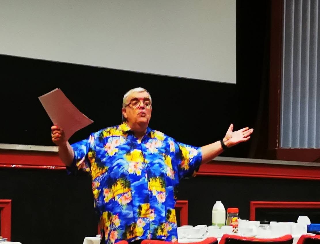 A man wearing a brightly colored Hawaiian shirt is standing in front of a presentation screen, gesturing with his arms outstretched, holding papers in his right hand.
