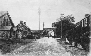 Black and white photo of a rural town street with houses on both sides and a bridge in the background.
