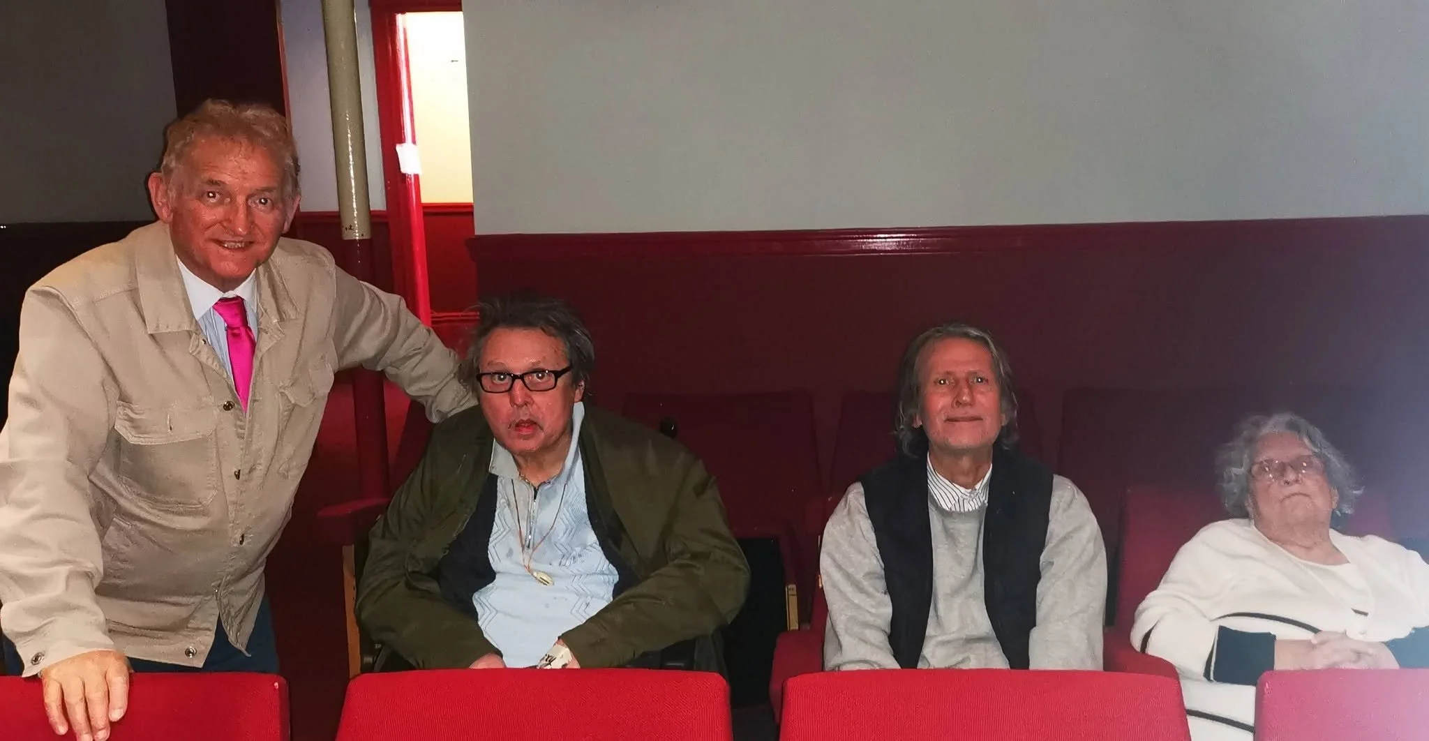 Four people sitting in a row in a room with red chairs and red walls, with one person standing on the left side. The seated individuals include a man with glasses and gray hair, a man with longer gray hair and a beard, a woman with glasses and curly 
