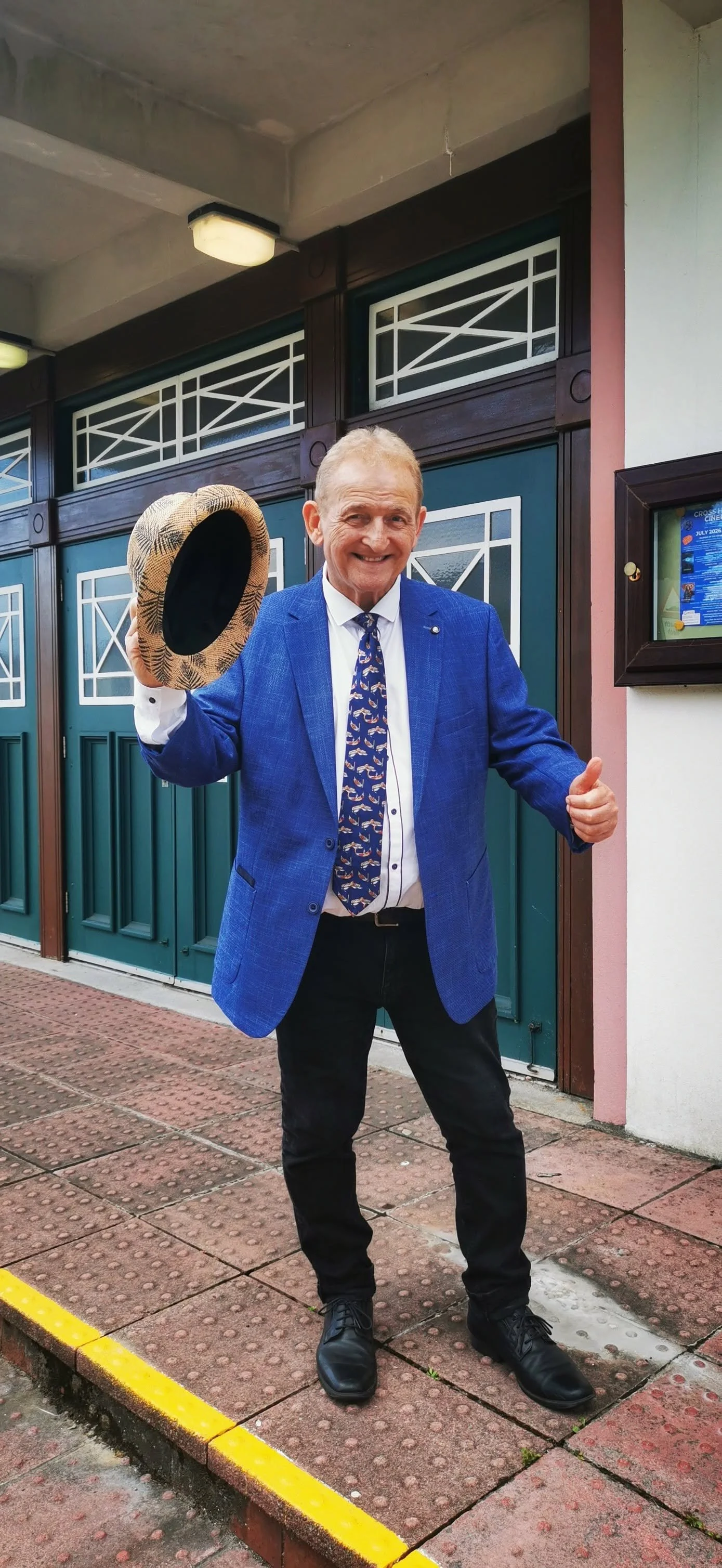 A man with gray hair, smiling, wearing a blue blazer, dress shirt, and a tie with a sailboat pattern, standing outside a building with green doors, holding a patterned hat, giving a thumbs-up.