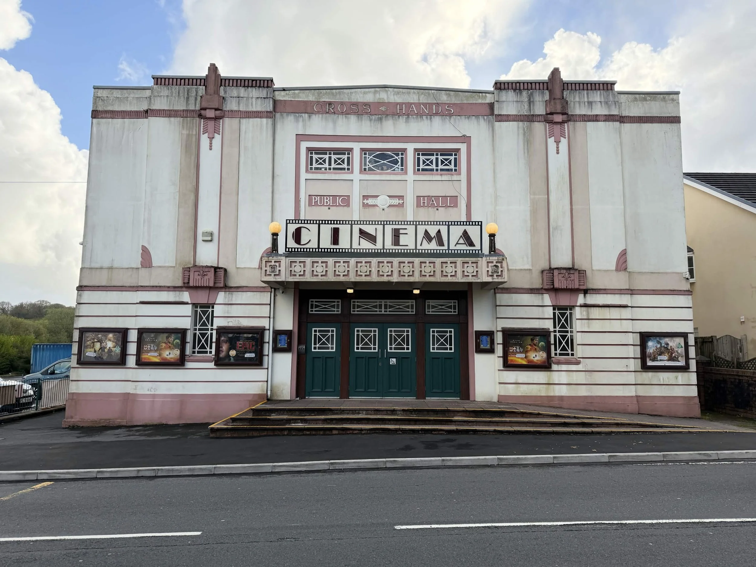 Facade of an Art Deco style building with a sign that reads 'CROSS HANDS' at the top and a marquee below that says 'CINEMA.' The building has decorative pink and cream-colored architectural details, and movie posters are displayed on the lower part of the facade. The sky above is partly cloudy.
