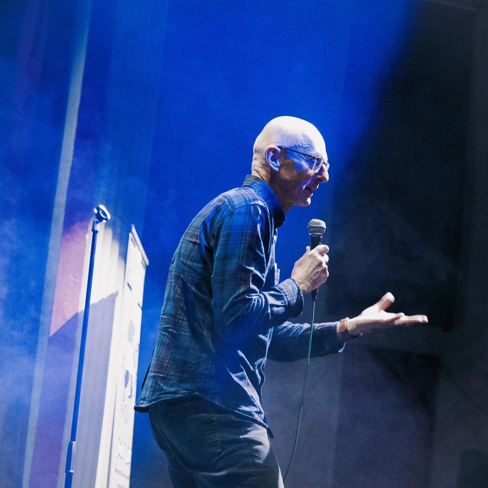 A man with glasses and a checkered shirt speaking or performing on stage with a microphone.
