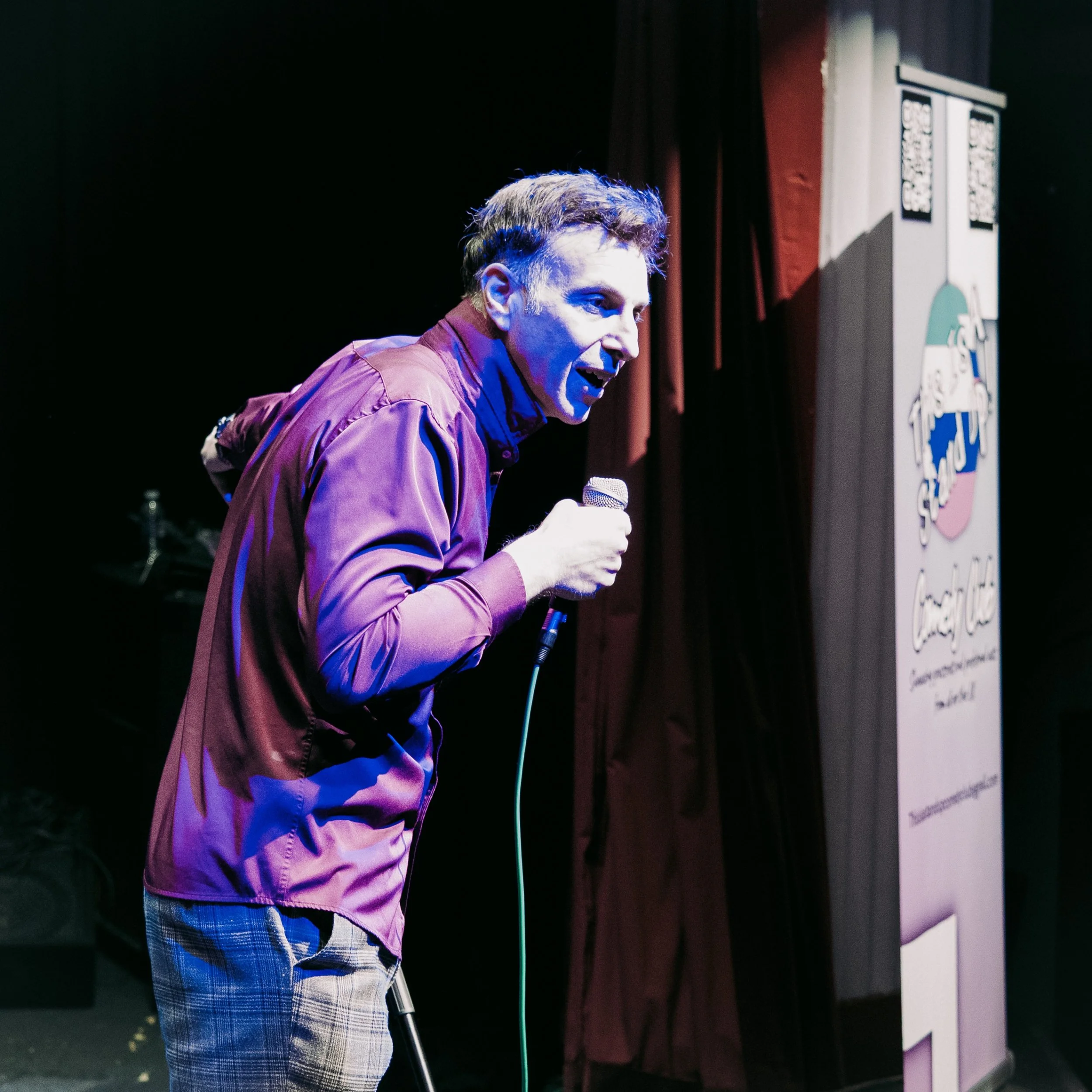 A man speaking into a microphone on stage, with a dark background and a sign partially visible on the right side.
