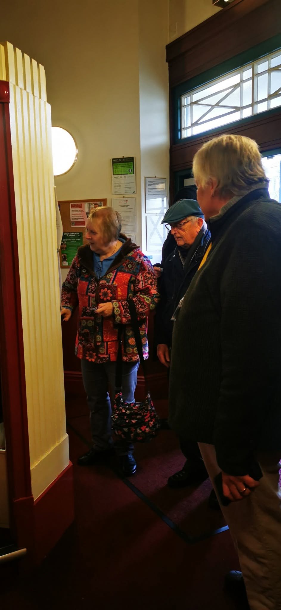 Three elderly people standing inside a building near a food counter, engaged in conversation.